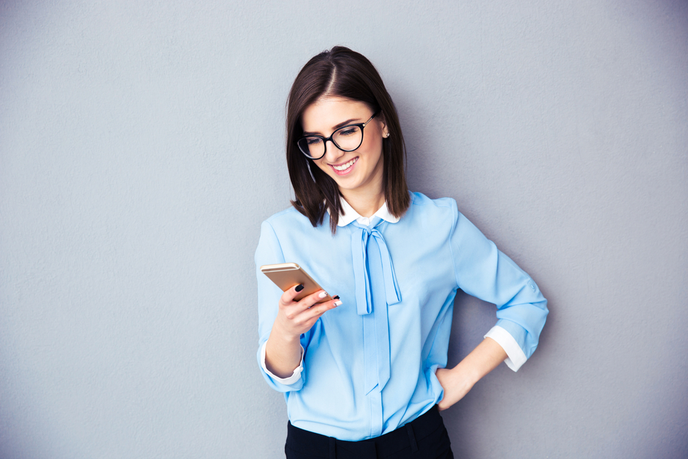 Smiling businesswoman using smartphone over gray background. Wearing in blue shirt and glasses.